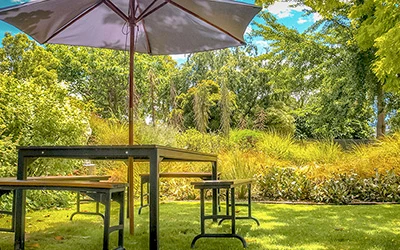 Grüner Garten mit vielen Büschen und Sitzbänken um einen Tisch mit großem Sonnenschirm